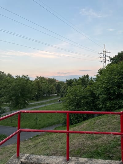 The image shows a park with a walking path, green trees, and a utility tower in the background. The sun is setting, casting a warm glow on the sky. A red railing is visible in the foreground. There are a few people walking in the park. The weather is pleasant. The scene appears to be in Oktyabr'skoe, Russia.