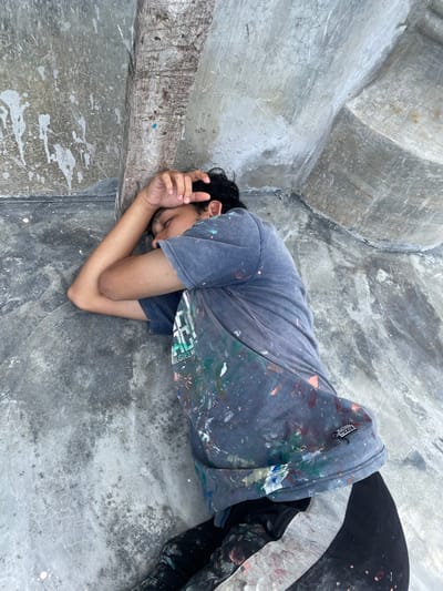 A person lies curled within a concrete structure. The individual rests their head on their hands, eyes closed. They wear a paint-splattered grey t-shirt and dark pants. The surrounding concrete is marked with streaks and spots of paint. The structure curves around the person, enclosing them within its gray, textured surface.