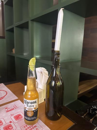 The image shows a still life arrangement of a dark green glass bottle with a white candle inserted into its neck and a bottle of Corona Extra beer with a lime wedge perched on its rim. The candle appears to be partially melted and dripping wax down the side of the bottle. The Corona bottle is a standard amber glass, and the beer inside is a pale yellow. The bottles are placed on a wooden table with a patterned paper placemat beneath the beer bottle. In the background, there are dark green shelves, possibly in a restaurant or bar setting.

The lighting suggests it is indoors, possibly during the day or evening. There are no people visible in the image. The scene appears to be staged, likely for a photograph, rather than capturing an ongoing activity. The visible text on the Corona bottle label includes "Corona Extra," "LA CERVEZA MAS FINA," and "BREWED UNDER LICENCE FROM CERVECERÍA MODELO, S. DE R.L. DE C.V." The placemat also has text in what appears to be Cyrillic script, with words like "BENTO," "ТОРТИК," "НАРОДЖЕННЯ," and "TAKE OUT." The overall mood is calm and still. The location context of Kryzhanivka, Ukraine, does not provide any immediately obvious visual cues within the image itself.