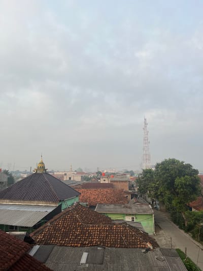 The image depicts a cityscape of Pasir Gadung, Indonesia, under a heavily overcast sky. The foreground is dominated by the roofs of residential buildings, characterized by varying materials such as clay tiles in shades of brown and grey corrugated metal. A prominent structure with a dark brown tiled roof features a distinctive gold and green domed top, adorned with a speaker or loudspeaker. Adjacent to this, another building exhibits a bright green facade.
In the midground, the urban landscape unfolds with more houses, some displaying orange tiled roofs. A tall, red and white lattice telecommunications tower stands prominently in the middle distance, equipped with multiple antennas. To the right of the tower, a large, leafy green tree partially obscures a paved road where a person on a bicycle is visible. Further in the background, several mosques are identifiable by their domes, including one with a green dome. The atmosphere suggests a hazy, possibly humid environment, with the distant buildings appearing softened by the atmospheric conditions. The overall scene captures a typical residential area within a developing urban setting in Indonesia.