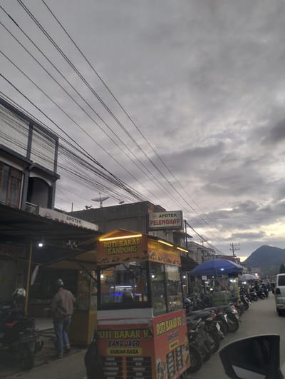 This is a medium-shot, vertical, outdoor, daytime, overcast photograph of a street scene in Gunungbukit, Indonesia. The sky is filled with heavy, gray clouds, suggesting impending rain or an overcast day. A street vendor's stall, advertising "ROTI BAKAR BANDUNG" (Grilled Bread Bandung) and "ROTI BAKAR RS BANG JAGO" (RS Bang Jago Grilled Bread) with a variety of flavors listed, is prominently featured in the foreground. The stall has bright yellow lights illuminating its signage and the products displayed inside.
To the left of the stall, a building with a sign that reads "APOTEK PELENGKAP" (Complete Pharmacy) is visible. Further down the street, more buildings and shops line the road, with a blue umbrella shading another vendor area. A row of parked motorcycles stretches along the side of the road, indicating a busy commercial area. In the background, a mountain is partially visible through the clouds, suggesting the mountainous terrain of the region.
A person in a light brown jacket and blue jeans is walking away from the camera near the roti bakar stall. Other individuals are partially visible in the background, possibly customers or vendors. The overall mood of the image is subdued due to the overcast weather, but the bright lights of the food stall offer a touch of vibrancy to the scene. The presence of the pharmacy and the food stalls suggests a lively local community.