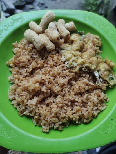 A close-up, overhead shot captures a bright green plastic plate filled with Indonesian cuisine. The plate is circular with an ornate raised pattern along the rim. The food itself consists of a generous portion of fried rice, interspersed with scattered spices and small pieces of protein, giving it a reddish-brown hue. To the right of the rice, a folded omelet, lightly browned and speckled with green herbs, rests on the plate. Several golden-brown, tube-shaped crackers, possibly krupuk, are arranged artfully on top of the fried rice. The background is blurred, showing natural elements like stones and foliage, suggesting an outdoor setting, possibly a backyard or a simple eatery in Cintadamai, Indonesia.