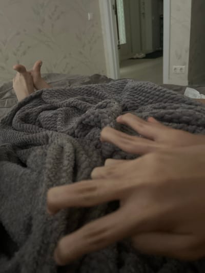 The image depicts a close-up view of two pairs of feet and hands under a grey, textured blanket, suggesting a domestic setting like a bedroom. The feet appear to be positioned near the top of the frame, with toes visible. In the foreground, two hands are present, one with fingers extended and the other partially obscured. The background shows a wall with patterned wallpaper and a doorway leading to another room, with furniture and a power outlet visible. The overall lighting is subdued. There is no indication of the specific location within Veshki, Russia.