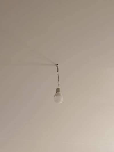 The image shows a simple ceiling with a single light bulb hanging from a wire. The light bulb is white and appears to be an LED bulb. The ceiling is a neutral color, possibly white or off-white. There is a shadow cast on the ceiling by the light bulb and its fixture. The image is taken from a low angle, looking up at the ceiling. The location is Izberbash, Russia.