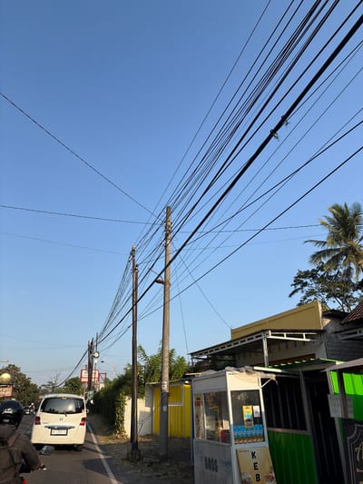 The image shows a street scene in the city of Kewaluhan, Indonesia, with a clear blue sky above. A series of overhead electrical wires dominate the upper portion of the frame, running diagonally across the sky and supported by wooden poles. In the foreground, a white car with an Indonesian license plate is traveling on the road, with a motorcycle rider behind it. To the right of the road, there are several small food stalls, one of which is labeled "BOSS KEBAB". Green and yellow structures are visible, and beyond them, trees and buildings can be seen. The overall impression is of a typical urban street in Indonesia, with a focus on the infrastructure of electrical cables and the presence of local businesses.