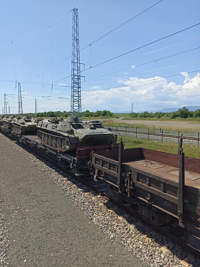 The image depicts a train carrying several armored fighting vehicles, likely BMPs, traveling on a railway line. The train is composed of flatbed cars and gondola cars. The surrounding environment features an open landscape with gravel ground, power lines supported by tall metal pylons, a fence, and some vegetation. In the background, there are hills and a partly cloudy sky. While not directly discernible from the image, the location is purportedly Zimnitsa, Bulgaria.