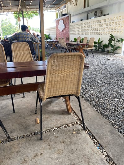 The image depicts an outdoor seating area of a cafe, potentially in Aluepiet, Indonesia. In the foreground, a woven rattan chair with a black metal frame sits on a concrete floor, partially obscuring a wooden table. Behind this table, another similar chair is visible. Further in the background, a man wearing a plaid shirt is seated at a table with other patrons, looking towards the left. The environment is characterized by a gravel ground covering, a white perforated wall with decorative patterns, and several potted plants. The cafe appears to be open-air, with a roof structure supported by poles. The lighting suggests it is daytime.