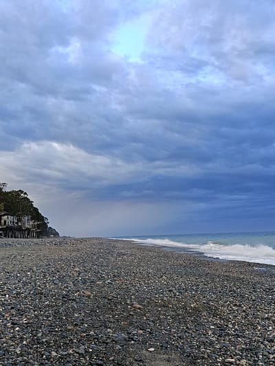 The image depicts a rocky beach in Gulrypsh, Abkhazia under a cloudy sky. The beach is covered in pebbles and stones, leading to a sea with small waves breaking along the shore. In the background, there are the remains of a decaying building complex, possibly a former resort or industrial structure, positioned on a small hill or raised area covered with trees. The sky is overcast with dark, heavy clouds, with a lighter patch visible directly overhead. No people are visible in the frame.