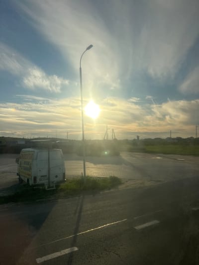 The image depicts a rural or suburban landscape with a white van parked on the left side of the frame. The van has markings on its side, including text that appears to be in Cyrillic. A street lamp stands near the center of the image, casting a long shadow across the asphalt road and a paved area behind the van. The sun is visible in the sky, creating a bright glare and illuminating wispy clouds. In the background, there are some industrial structures or buildings, power lines, and a line of trees or vegetation, suggesting a semi-industrial or undeveloped area. The overall scene appears to be set during the late afternoon or early evening, given the low angle of the sun and the lighting conditions. The location is identified as Kamianopil, Ukraine.
