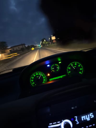 In Mezzana Sant'Antonio, Italy, at night, the image captures a view from inside a car, looking forward through the windshield. The dashboard displays illuminated green gauges, with the speedometer indicating a speed of approximately 100 km/h. The digital display below the gauges shows "Mar. 15. 51." The headlights of the car illuminate the road, which is lined with trees and some buildings on the left. The sky is dark and cloudy, suggesting a potentially stormy evening. The car's radio is also visible, displaying "3 MUS" in illuminated text. No individuals are visible in the image.