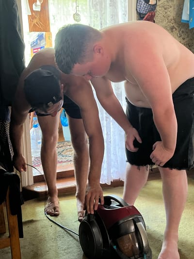 In a dimly lit room, possibly a home in Sad. Uchastki, Russia, two young men are engaged in cleaning. One man, wearing a black baseball cap and black shorts, is bent over and appears to be operating a red and black LG vacuum cleaner on a worn, patterned carpet. The other man, shirtless and also wearing black shorts, is positioned behind him, looking down at the vacuum cleaner with his hands near the machine, as if assisting or observing. The environment appears cluttered, with a doorway in the background revealing more of the interior. Sunlight filters through a sheer curtain, casting a soft glow on the scene. There are two large plastic water bottles visible near the doorway. The overall impression is one of an ordinary domestic task taking place in a rustic setting.