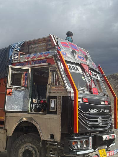 In Bodhkharboo, India, a heavily decorated Ashok Leyland truck is parked under a dramatic, cloudy sky. The truck features intricate colorful designs and lettering on its exterior, with one inscription reading "JANNAT E KASHMIR". A young person, dressed in a teal-colored garment and a scarf, is seated on the roof of the truck, looking towards the right. The truck is a dark color with prominent chrome accents and red support bars on the front. The background shows a mountainous landscape with dry, rocky terrain. The scene appears to be a roadside stop, possibly during travel.