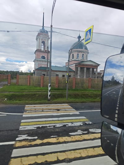 A side-view mirror of a vehicle reflects a rural road and sky as the vehicle approaches a pedestrian crossing. In the background, a pink and white church with a green dome and a bell tower stands behind a wooden fence. The sky is overcast with some patches of blue.