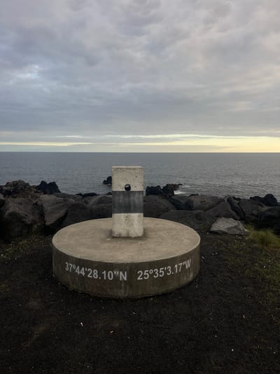The image depicts a concrete marker with geographic coordinates against a backdrop of a rocky coastline and ocean. The marker is cylindrical with a square pillar on top, painted with horizontal black and white stripes. The coordinates "37°44'28.10"N 25°35'3.17"W" are inscribed on the cylindrical base. The background includes dark, volcanic rocks meeting the ocean water, and a cloudy sky above. The ground surrounding the marker is black, volcanic soil. The location is Lagoa (Nossa Senhora do Rosário), Portugal. No people are visible.