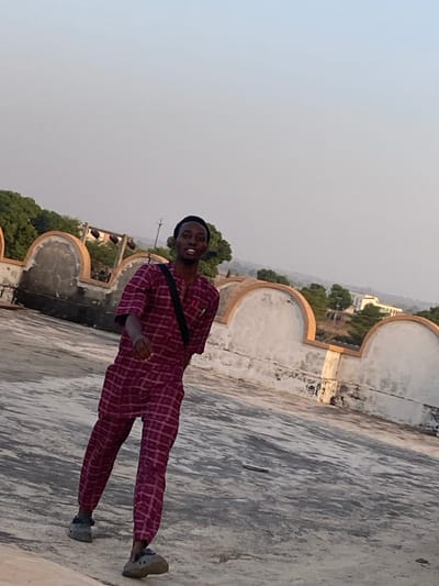 A single dark-skinned adult male is depicted in Barako, Nigeria, standing on a weathered, light-grey concrete rooftop. He is captured in mid-stride, with his right foot slightly raised, and his body angled towards the right while his head is turned to face the camera. He is smiling, with teeth visible. He wears a black beanie, a long-sleeved collared shirt, and matching trousers, all featuring a maroon and white plaid pattern. A black strap or bag is worn diagonally across his chest. His feet are clad in grey, possibly Crocs-style, clogs. Behind the man is a low, light-colored wall with several decorative arched sections, showing signs of wear and discoloration. Beyond this wall, the background reveals trees and a distant light-colored building under a hazy, bright sky.