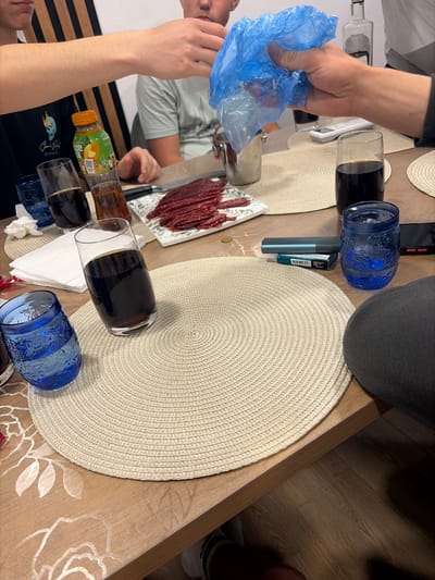 The image captures a casual indoor gathering around a wooden table in Dolno Sahrane, Bulgaria. Several individuals are partially visible, suggesting a social event. A prominent hand holds a blue plastic bag, possibly in the act of preparing or distributing food. Another person, wearing a light green shirt, is seen reflected in the table's surface, while a third person in the foreground wears a dark t-shirt with "Grand Prix" visible text and graphics.
The table is set with multiple round, light beige woven placemats on a tablecloth featuring a subtle white floral pattern. Various beverages are present: several clear glasses filled with a dark liquid (likely beer or a dark soda), two ornate blue glasses, an orange "FRUCTIS GRAPEFRUIT 100%" juice bottle, and a clear bottle that could contain water or spirits. A white rectangular plate holds numerous slices of dried or cured meat, possibly sausage.
Other objects on the table include white napkins, a black smartphone, a blue vaping device, and a pack of "HEETS" tobacco sticks. A small gold-colored coin rests near one of the placemats. The indoor lighting suggests an evening or nighttime setting. The floor visible in one section is made of light wooden planks. The overall atmosphere appears informal and relaxed.