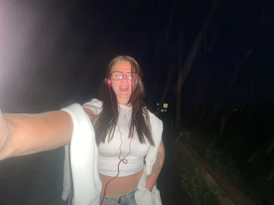 A nighttime photograph, taken in Molokovo, Russia, shows a woman wearing glasses and headphones, hanging upside down. The woman is wearing a white tank top and a white cardigan, with a glimpse of patterned pants. Her hair is dark brown, and her mouth is open. A forearm is prominently displayed at the top right of the frame. The background includes dark foliage and dim streetlights.