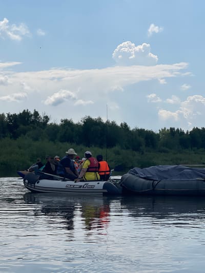 The image shows an inflatable boat with several people on board, navigating a body of water. The boat is white with black trim and has the brand "POLAR BIRD" and model "340 M" visible on its side. People are wearing life jackets in various colors, including yellow, orange, and blue. Some are wearing hats. The boat is moving away from the viewer, towards a dense treeline on the opposite bank of the water. The sky above is partially cloudy with a light blue hue. The water reflects the sky and the treeline. Another, larger inflatable object, covered with a gray tarp, is tied to the side of the main boat. The scene appears to be a recreational outing on a river or lake. The location is identified as Nezhinka, Russia, suggesting a natural environment.