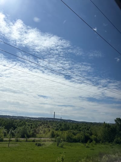 The image displays an expansive outdoor scene in Vyatskiye Polyany, Russia, under a bright, partly cloudy sky. The upper portion of the frame shows a clear blue sky with widespread altocumulus clouds, denser on the left and more broken towards the right. A bright sun reflection or lens flare is visible in the upper left corner. Three dark, thin power lines run diagonally across the sky from the upper left to the lower right. The lower half of the image features a lush, green landscape. The foreground consists of overgrown grassy terrain. In the midground, a dense band of deciduous trees and bushes stretches horizontally, partially obscuring a narrow, unpaved road or dirt path. Several dark utility poles stand upright near the path within the vegetated area. On the distant horizon, a significant industrial complex is visible, characterized by multiple large buildings, storage tanks, and at least two tall, slender smokestacks or chimneys, one distinctly dark and prominent, suggesting industrial facilities. No people, animals, or vehicles are present in the scene.