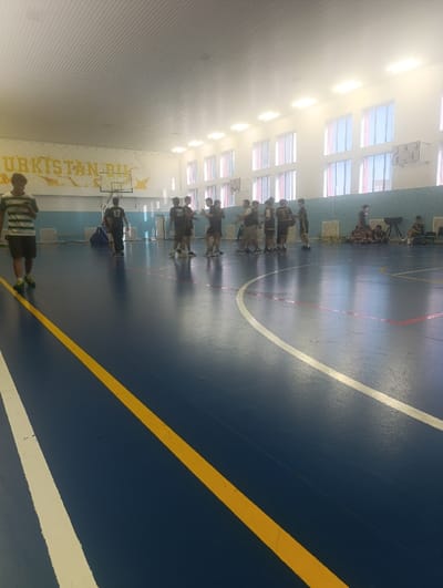 Inside a brightly lit gymnasium, a group of people gathers on a blue basketball court with yellow and white lines. Some individuals are in athletic wear, possibly basketball players, while others sit along the wall. A basketball hoop stands against the wall, which is decorated with the word "UZBEKISTAN." Windows line the upper portion of the wall, letting in natural light.