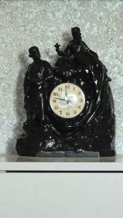 The image shows a black, ornate clock with a round face featuring Arabic numerals and two hands. The clock is mounted on a sculpted base that depicts two figures, possibly a man and a woman, in a standing posture. The background is a textured, light-colored surface that appears to be wallpaper with a shimmering or glittery effect. The clock and its base are positioned on a white surface, possibly a shelf or table, against this textured wall. The overall scene is an interior shot. There is no visible indication of the city of Neftegorsk, Russia.