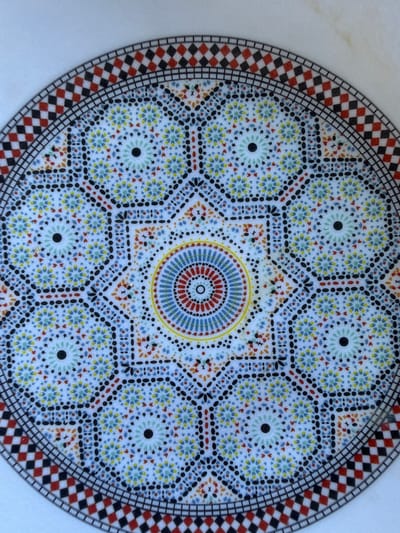 An overhead, close-up shot captures a decorative, circular object, likely a ceramic plate or a mosaic tabletop, set against a plain white background. The object showcases an intricate, symmetrical geometric pattern typical of Moroccan artistry.
At the very center, an eight-pointed star is formed, enclosing concentric rings of small red, blue, yellow, and black dots, with a small open circle at its core. Surrounding this central star are eight smaller, circular motifs resembling stylized flowers or stars. Each of these smaller patterns features a prominent black dot in its center and is filled with tiny dots in shades of light blue, yellow, red, and black. The spaces between these elements are filled with complex geometric designs comprising black, red, and orange dots, forming additional star-like and hexagonal shapes.
The outermost edge of the circular design is a wide border made up of alternating red and black checkered squares, simulating a mosaic tile pattern. This border is delineated by several thin, concentric black lines. The overall color palette is vibrant, utilizing various shades of blue, red, yellow, orange, and black, all set against a white base. The intricate detail and geometric precision reflect the rich cultural heritage associated with the specified location of Loudaya, Morocco. There are no people, visible activities, or text in the image.