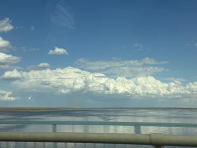 This is a landscape photograph taken from a moving vehicle, likely a car, as indicated by the presence of a railing in the foreground that appears to be part of a bridge or overpass. The scene depicts a wide expanse of calm, blue-grey water under a bright, partly cloudy sky. The horizon is flat, with a low, dark strip of land visible in the distance, suggesting a coastal or deltaic environment. Scattered white birds are visible on the water.

The sky is dominated by a vibrant blue, with fluffy white cumulus clouds in the middle ground and less defined, wispy clouds higher up. There are also some unusual bright, lens flare-like reflections in the sky, possibly caused by the sun or light sources outside the frame. The overall impression is of a clear and sunny day, likely during daylight hours. The water is smooth and reflects the sky and clouds, creating a serene and expansive feeling. The railing in the foreground, painted in muted blue and grey tones, creates a sense of depth and places the viewer within the scene. No people or specific man-made objects other than the railing are clearly identifiable. The environment suggests a natural, open setting.