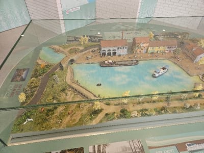 This image captures a detailed miniature diorama, depicting a bustling canal basin scene in the city of Avignonet-Lauragais, France. The central feature is a light blue, irregular-shaped water body representing a canal, with two primary miniature boats: a dark brown barge-style vessel docked near the left bank and a white leisure boat with a cabin positioned further out in the basin. A smaller dark rowboat is also present near the far bank.
On the left bank, a cluster of miniature multi-story buildings with tiled roofs lines the water's edge, including a prominent grey-stone structure with multiple arched openings and an industrial chimney visible in the background. The right bank is populated by several two-story buildings in pastel colors such as yellow, light blue, and orange.
The surrounding landscape features numerous miniature deciduous trees and shrubs in shades of green and yellow, distributed across the banks and terrain. Dirt paths and unpaved roads crisscross the model. A miniature stone arch bridge spans a narrower waterway on the far left. Numerous miniature human figures are scattered throughout, depicted walking along the canal paths, congregating near buildings, and appearing on the bridge. Several miniature brown cows are visible grazing in a grassy field in the lower-left section. The entire diorama is enclosed under a clear glass panel, reflecting ambient light, and surrounded by informational graphics and text, including historical images and text referencing a "PONT EN DOS D'ANE" (donkey-back bridge), providing context about the Canal du Midi region.