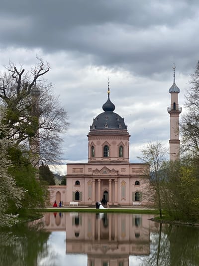 The image depicts a building with a prominent dome and a minaret-like tower reflected in a body of water, likely a pond or lake. The building is light pink or beige in color, with ornate architectural details. The dome is dark gray with a gold-colored crescent-shaped finial on top. The tower is cylindrical and has a narrower section at its top with a similar finial.

The surrounding environment includes bare trees and some budding greenery, suggesting early spring or late autumn. People can be seen walking along the bank of the water near the building. The sky is overcast with gray clouds. The reflection in the water is mostly clear.

The location is Schwetzingen, Germany, specifically the Schwetzingen Palace gardens, where the mosque is located.