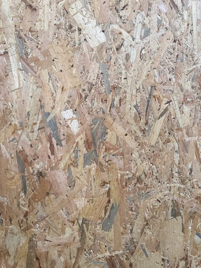 The image is a close-up, overhead shot of a particleboard surface, likely an Oriented Strand Board (OSB). The texture is rough and uneven, composed of compressed wood chips and strands of varying sizes and colors, ranging from light tan to darker brown and gray. The light casts minimal shadows, suggesting a diffuse or overhead light source. There are no discernible objects, people, or actions within the frame. The scene is devoid of any recognizable urban or natural environment, and therefore, its location in Fontevivo, Italy, cannot be determined from the image itself.