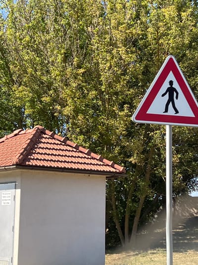 In Tvrdonice, Czechia, a triangular red and white pedestrian crossing sign stands by a pole. The sign depicts a black silhouette of a person wearing a hat and walking. To the left, a small, light-colored building with a red tile roof is visible. The building has a metal door on its left side, with some text printed on it. Behind the building and sign, lush green trees form a dense backdrop. The ground in front of the building and sign is covered in grass and dry leaves, with some dirt visible.