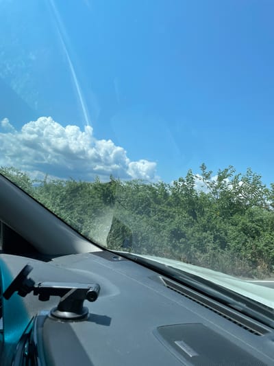The image is taken from inside a vehicle, looking out through the windshield on a sunny day. The dashboard of the vehicle is visible in the foreground, with a black suction cup mount for a device attached to it. Beyond the dashboard, a dense line of green trees and vegetation stretches across the middle ground. The background is dominated by a bright blue sky with scattered white, puffy clouds. The scene appears to be in a rural or semi-rural area. No people or specific landmarks are visible to pinpoint a location within Tsurtavi, Georgia, but the presence of lush vegetation suggests a location with sufficient rainfall and greenery.