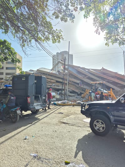 The image depicts a scene of destruction in Caraballeda, Venezuela, following a catastrophic event, likely a landslide or earthquake, given the context of past natural disasters in the region. A collapsed multi-story building, its concrete structure shattered and debris strewn across the area, dominates the background. Yellow construction equipment, specifically a backhoe loader, is present, suggesting ongoing or imminent rescue and recovery efforts.

In the foreground, authorities are present, indicating a response to the disaster. A dark, militarized van with the number "062" visible on its rear is parked on the street. A person in a red shirt and hard hat is walking near the van, possibly involved in the rescue operation. Another individual, wearing a vest with "MPPH" visible, is seated on a motorcycle to the left of the van, also seemingly involved in the scene's management. A second dark SUV is parked in the lower right portion of the image, its front wheel and bumper visible. Scattered debris, including a plastic bottle and what appears to be trash, litters the asphalt street. Overhead, power lines are visible, strung between poles, with tree branches extending into the frame from the left. The sun is bright, casting strong shadows.