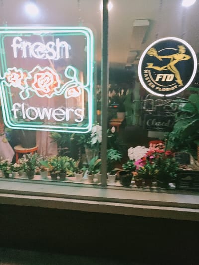 The image shows a storefront window of a florist located in Richmond, Virginia, at 37.548 latitude and -77.463 longitude. The window displays a neon sign that says "fresh flowers" with stylised roses in the middle. There is another circular sign with a logo of a running man and the words "FTD Master Florist". The window also displays several potted plants. The store appears to be closed as a sign that says "Closed" can be seen behind the potted plants. The shop is illuminated from inside with light spilling out from a storefront window. A light fixture is visible above the window. The building facade is brick. The image was captured during nighttime.