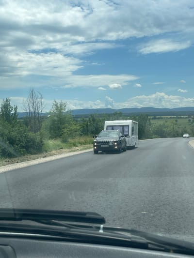 The image shows a view from inside a vehicle on a road near Probuda, Bulgaria. In the distance, there are slightly rolling, tree-covered hills. A black car is overtaking a white caravan on a two-lane asphalt road. The sky is light blue with scattered clouds. The camera is looking out of the windshield from within the vehicle.