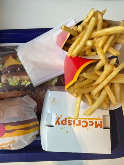 An overhead shot captures a McDonald's fast-food meal arranged on a blue plastic tray. Two containers of golden-brown french fries are prominent; one is in the iconic red and yellow McDonald's paper carton, and the other appears to be in a plain white paper container. A white cardboard box, labeled "McCrispy" in orange letters with scattered orange dots, rests on the tray, showing slight grease stains. Below it, a burger is partially visible, wrapped in white paper with yellow and brown stripes, and the text "Doub Cheeseb" (likely "Double Cheeseburger") is visible on its side. A white paper napkin is tucked under one of the fry containers. The tray liner, or the surface beneath, features a large graphic of a burger. The scene suggests a meal recently purchased or about to be eaten at a McDonald's restaurant. The provided context places the location in Chuchuligovo, Bulgaria.