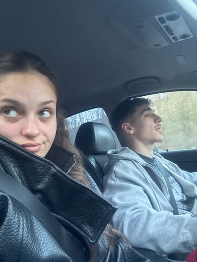 The image depicts the interior of a vehicle during daylight in Gorna Vasilitsa, Bulgaria. Two young adult individuals are seated in the front seats. A female occupies the front passenger seat, positioned in the foreground and to the left. She has long brown hair and is wearing a black textured leather-like jacket with a visible seatbelt crossing her chest. Her eyes are directed upwards and to her right, away from the camera, and her lips show a subtle smirk. A male occupies the driver's seat, positioned in the background and to the right. He has short dark hair with a faded haircut and light facial hair. He is wearing a light grey zippered hooded sweatshirt over a dark t-shirt, also with a visible seatbelt. His head is turned slightly to the right, and his gaze is directed forward and out of the vehicle. The car interior features dark trim, a dark headliner, an overhead console, and a grab handle above the passenger window. Through the windows, a blurred outdoor environment is visible, consistent with trees or vegetation alongside a road.