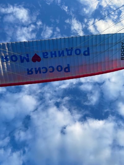 The image shows a paraglider with Russian text and a heart symbol against a blue sky with clouds. The text on the paraglider, when read from right to left and upside down, says "РОДИНА МОЯ РОССИЯ" which translates to "My Motherland Russia". The paraglider has a red trim along its bottom edge and the brand name "ROCKY" is visible vertically on the right side. The paraglider is suspended in the air by lines, suggesting it is either in flight or about to be launched. The environment is an outdoor setting with a clear blue sky and scattered white clouds. The location is specified as Makarovka, Russia. There are no people or specific actions clearly depicted in the image.