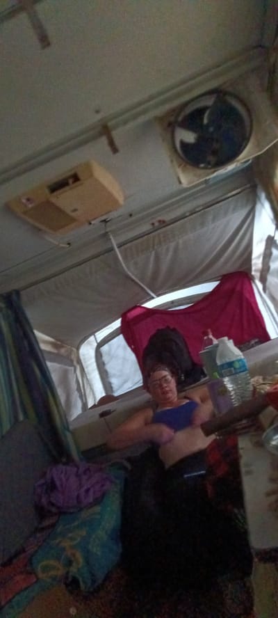 The image shows the interior of a pop-up camper, possibly located in Clarksville, United States of America. A woman wearing glasses and a blue top is lying down inside. A red shirt is hanging from the camper's roof support. On the ceiling are an air conditioning unit and a fan. Other objects include a water bottle, a cup, and various fabrics/blankets.