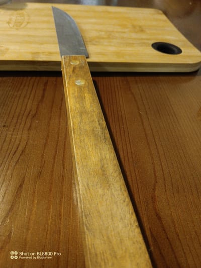 The image shows a close-up view of a wooden-handled knife lying on a wooden cutting board, which is itself on a wooden table. The knife's blade is made of metal and is positioned diagonally across the cutting board. The handle is made of light-colored wood and is attached to the blade with two visible rivets. The cutting board is light brown and has a dark circular cutout on the right side. The table surface is a darker brown with visible wood grain. There are no people or any other objects present in the scene. The image appears to be taken indoors, likely in a kitchen setting. The bottom left corner of the image contains a watermark that reads "Shot on BL8800 Pro Powered by Blackview". There is no information available in the image to suggest a specific location in Durankulak, Bulgaria.