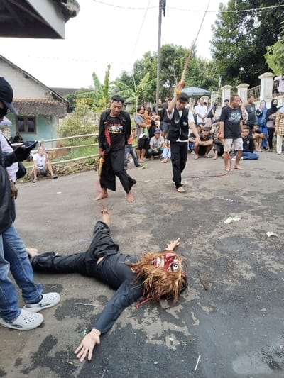 The scene depicts an outdoor event on a paved road in Sumberejo, Indonesia, observed by a crowd. In the foreground, a figure lies supine on the dark asphalt, wearing black long-sleeved attire and pants, with bare feet. The figure's head is covered by a large, ornate mask featuring a red face, wide white eyes, prominent fangs, and long, reddish-brown hair. Red string-like material is visible on the ground near the figure's bare feet.

Standing over the prone figure, slightly to the left, is a barefoot male wearing a black long-sleeved shirt with a red and yellow design, black pants, and a red headband. He holds a short, curved wooden implement in his right hand. Directly to his right, another barefoot male walks forward, wearing a white t-shirt under a black vest with white patches, and black pants. He holds a long, thin staff or pole with a decorative element at its tip, raised above his head. Further to the right, a third barefoot male stands, dressed in a black t-shirt and light shorts, holding a red braided rope or whip.

The background features numerous spectators, adults and children, gathered along the sides of the road, some standing, some crouching. Houses with tiled roofs and a variety of green vegetation, including trees and banana plants, are visible beyond the crowd. A utility pole stands mid-ground on the left. The sky is overcast.