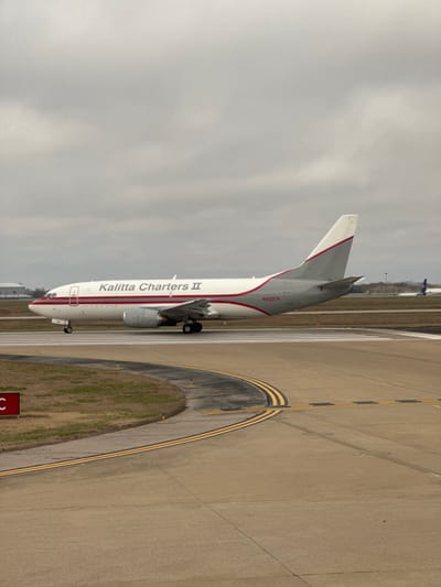 A white and grey Kalitta Charters II Boeing 737-type cargo aircraft, registered N332CK, is positioned on a concrete airport taxiway at Nashville International Airport (BNA) in Nashville, Tennessee, United States. The aircraft's fuselage is white with a prominent red stripe running horizontally, while its tail section is grey with a white top and a diagonal red stripe. Two turbofan engines are mounted under the wings. The foreground shows a curved section of concrete taxiway marked with double yellow lines, indicating taxiway edges, and white lines for runway hold positions. Adjacent to the taxiway on the left is a patch of dormant, brown-green grass with a red square sign displaying a partial white letter 'C'. The sky is uniformly overcast and grey. In the distant background, faint structures consistent with airport infrastructure and the purple tail of another aircraft are visible. No people are present in the scene.