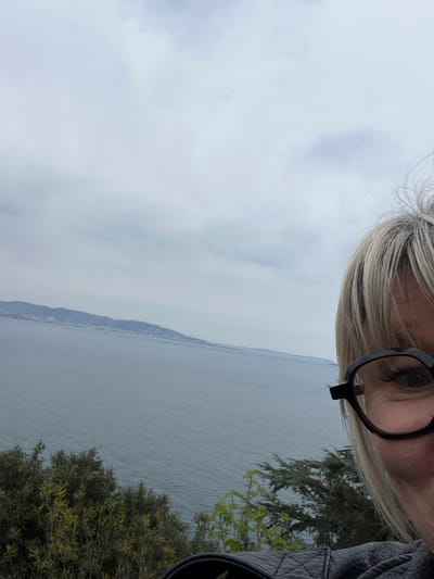 The image captures an elevated coastal view overlooking the Mediterranean Sea near Théoule-sur-Mer, France, under an overcast, light grey sky. A person's partial face and upper body are visible in the lower right, featuring a right eye with visible eyelashes behind square, dark-framed glasses. Blonde hair frames the face, and a black quilted leather jacket is visible on the right shoulder and arm. The expansive, rippled grey-blue sea occupies the midground, with a distant, hazy coastline featuring hills and scattered buildings visible on the horizon. Dense green and yellowish-green foliage, including shrubs and trees, occupies the immediate foreground, suggesting an elevated vantage point.