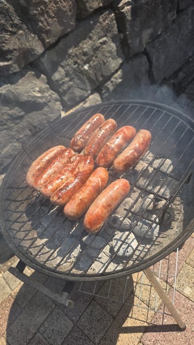 A high-angle shot captures several sausages grilling on a charcoal barbecue in Albstadt, Germany. The barbecue is circular with a black metal grate over grey charcoal. Eight sausages are visible on the grate, some appearing slightly browned and charred. In the background, a rough stone wall forms the backdrop, with sunlight casting shadows. The ground beneath the barbecue is paved with interlocking concrete tiles. The scene suggests an outdoor cooking activity, possibly in a backyard or patio area within the city.