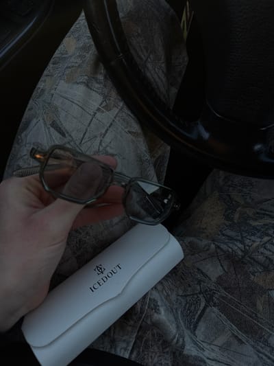 The image depicts an indoor scene, likely inside a vehicle, given the partial view of a black steering wheel on the far left. The setting is characterized by a camouflage-patterned fabric covering what appears to be a car seat, featuring muted tones of grey, brown, and black, reminiscent of tree bark or autumn leaves.

The central subjects include a person's hand, a pair of eyeglasses, and an eyeglasses case. A fair-skinned hand is positioned in the lower-center, gently holding a pair of modern eyeglasses with dark, possibly translucent, frames. To the right of the hand, a white, rectangular eyeglasses case rests on the patterned seat fabric.

The activity taking place is simple: a person is handling their eyeglasses, possibly in the process of putting them on, taking them off, or preparing to store them in or retrieve them from the nearby case.

Notable details include the dim and soft lighting, which suggests either an indoor environment with limited natural light or an early morning/late evening time. There are no explicit emotional cues. The most prominent visible text is on the white eyeglasses case, which clearly displays the word "ICEDOUT" in black uppercase letters, accompanied by a stylized black emblem or logo resembling an ornate letter 'E' positioned above the text. The image itself does not contain specific visual cues to identify the location as Chargan, Bulgaria, beyond the context provided.