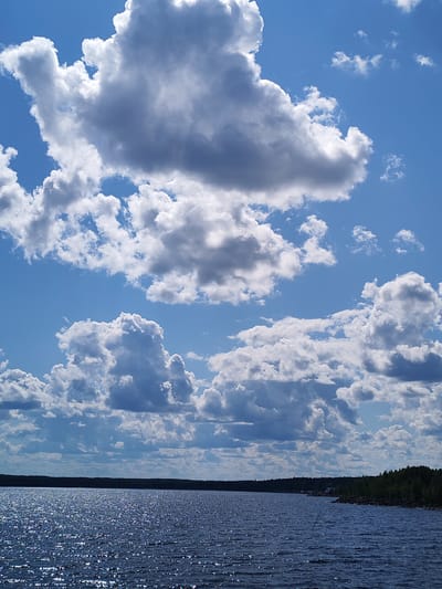 The image depicts a wide expanse of dark, rippling water under a bright blue sky filled with dramatic white and gray cumulus clouds. The water's surface is textured with countless glints of sunlight. Along the horizon, a dense line of dark green trees forms a shoreline, with a darker, indistinct silhouette of land beyond. The overall scene suggests a natural, possibly remote, location. No people or specific man-made structures are clearly discernible within the frame, though a very faint hint of a distant building or structure might be present on the far right edge of the treeline. The location is identified as Kvartsitny, Russia.