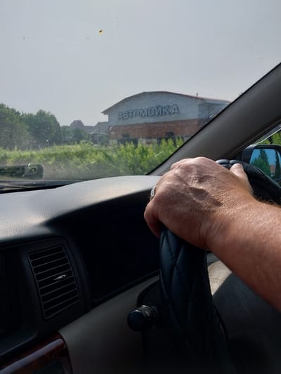 The image shows a first-person perspective from inside a car, with a hand on the steering wheel. The hand is lightly gripping the wheel, suggesting the car is in motion or about to be. The dashboard and a portion of the passenger side are visible. Outside the car, through the windshield, a building with the word "АВТОМОЙКА" (Car Wash) in large letters is visible in the distance. The building appears to be a commercial establishment. The surrounding environment includes trees and greenery, indicating an outdoor setting. The sky is bright and appears hazy, possibly due to sunlight or atmospheric conditions. The scene is likely set in a suburban or semi-rural area. The presence of the "Car Wash" sign strongly suggests the location is within a town or city, and given the Cyrillic script, it is in a Russian-speaking region. Belokurikha, Russia, is a known resort town with numerous car washes serving tourists and locals.
