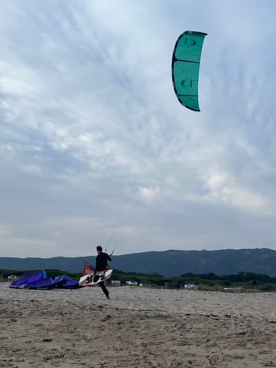 A kite surfer, dressed in black, carries his board and is preparing to enter the water on a sandy beach. He is moving from left to right across the frame. A large, teal-colored kite is visible in the sky, connected to him by lines. In the background, several other kites are laid out on the sand, along with a dark, mountainous landscape. The sky is overcast with clouds, typical of the windy conditions Tarifa is known for.