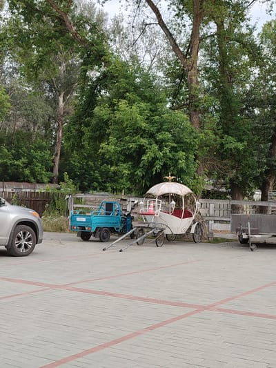 A light-colored car is parked in a lot with a light gray paver surface marked with red lines. To the right of the car is a small blue truck. Further to the right is a white carriage with a decorative canopy and red seating. In the background, there is a wooden fence and numerous trees. The location is identified as Shcherbani, Ukraine.
