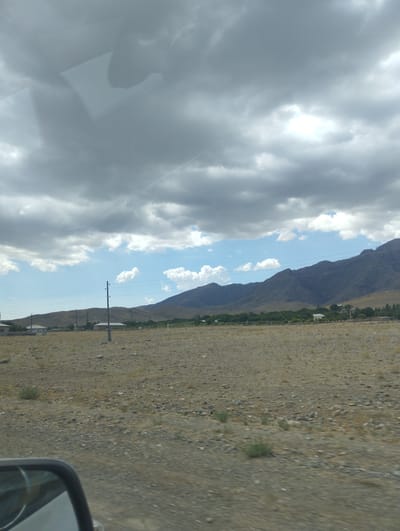 The image depicts a view from a vehicle, looking out at a rural landscape under a cloudy sky. In the foreground, a side-view mirror is partially visible on the left. The ground is covered with gravel and sparse, dry vegetation. In the middle ground, there are a few low-lying buildings with simple roofs, interspersed with trees. A solitary utility pole stands prominently in the field. In the background, a range of low, barren mountains stretches across the horizon. The sky is filled with a mix of dark, heavy clouds and patches of brighter sky, suggesting a partly cloudy or overcast day. The scene is likely located in a rural area of O'xum QFY, Uzbekistan, characterized by its arid terrain and mountainous backdrop. There are no people visible in the image.